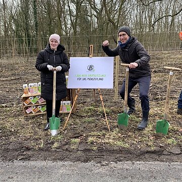 9100 Bäume für Beckumer Wald Polaroid Zwei Personen mit Schaufeln vor einem Wald mit einem Aufsteller mit einem Berief-Plakat, im Hintergrund Berief Getränkekartons.