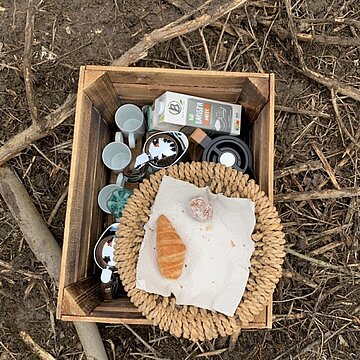 Über 9100 Bäume für Beckumer Wald Polaroid Ein kleiner Holzkasten in dem ein Berief Getränkekarton zusammen mit Tassen und Kannen, darauf ein Korb mit einem Croissant auf einem Waldboden.