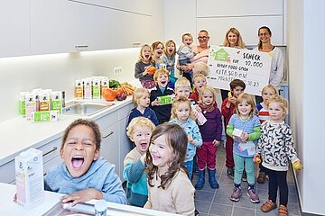 Sieger der Aktion grüner Kindergarten Kinder des Kindergartens lachen in die Kamera, halten ein Plakat von Berief hoch, daneben Getränkekartons von Berief.