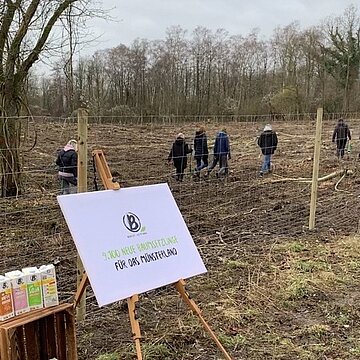 Über 9100 Bäume für Beckumer Wald Polaroid Ein Berief Plakat auf einem Aufsteller vor einem Maschendrahtzaun auf einer Brache, dahinter gehen Menschen umher.