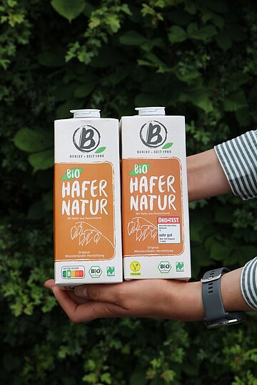 Ohne Nutriscore Zwei Hafergetränkekartons werden vor einem Blätterhintergrund in die Kamera gehalten.