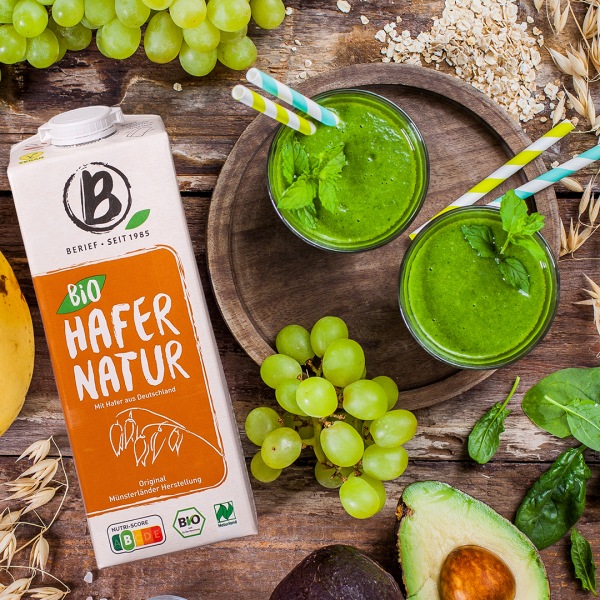 Haferdrink alles über unseren Hafer Drink Karton Hafer-Drink neben einem Holzbrett auf dem zwei grüne Smoothies stehen mit Früchte-Dekoration