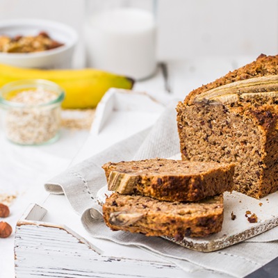 Saftiges Bananenbrot a la Berief Bio Hafer Drink Angeschnittenes Bananenbrot auf einem weißen Tischtuch mit Dekoration