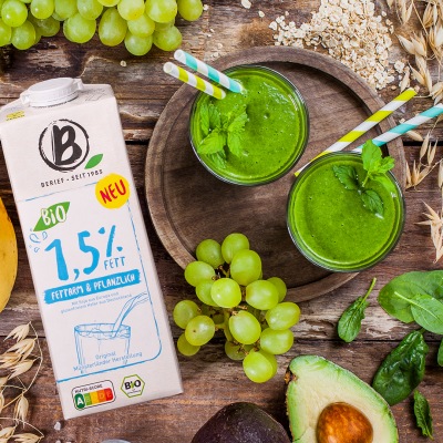 Grüner Smoothie mit Spinat und Avocado 2 grüne Smoothies in Gläsern mit Strohhalm auf einem Holzbrett mit Getränkekarton.